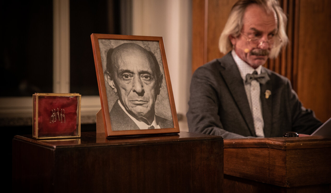 Auf dem Foto ist ein Bild eines in Warschau ermorderten, älteren Mannes zu sehen. Daneben leitet ein Dirigent das Orchester.