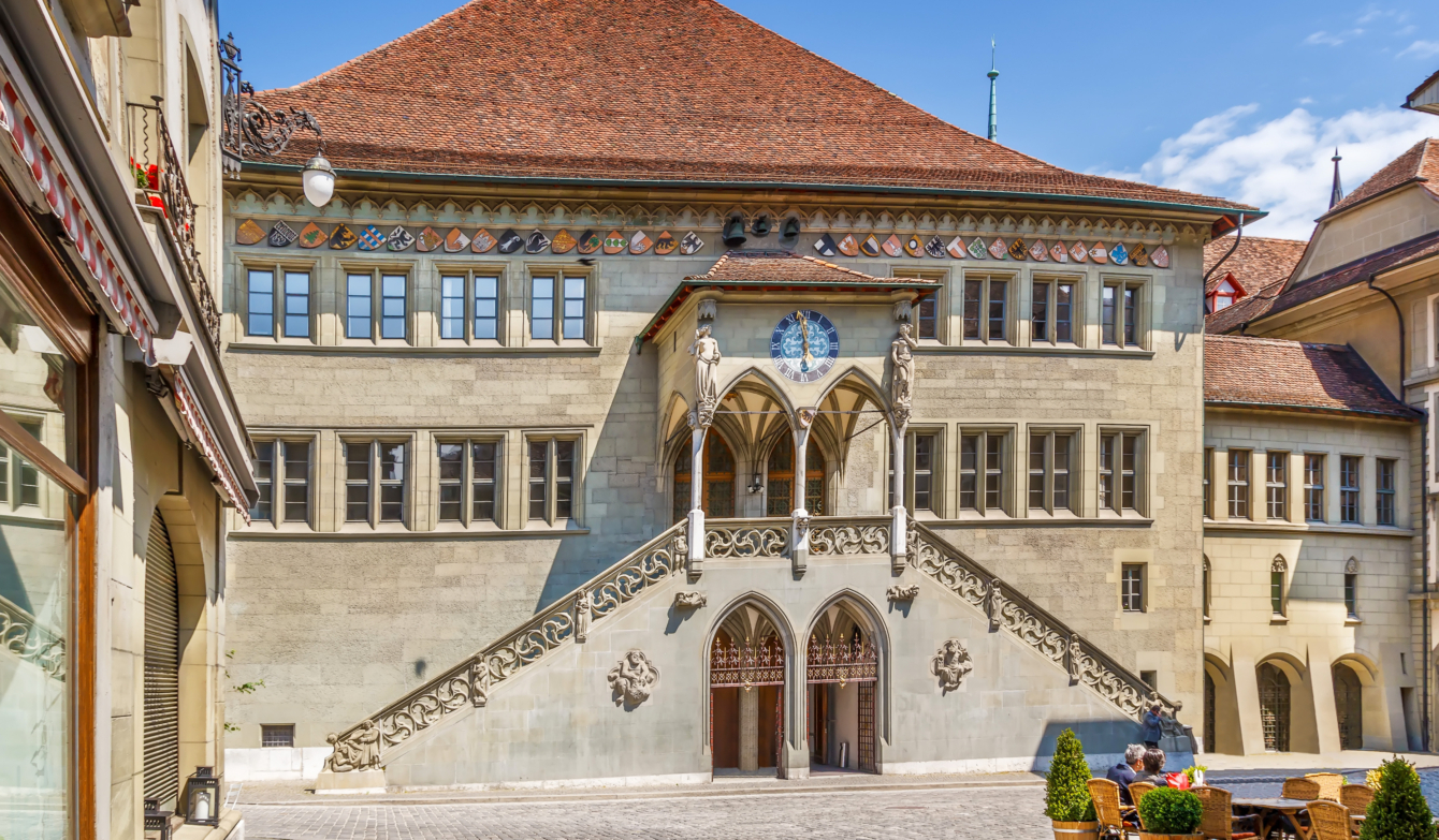 Berner Rathaus von vorne fotografiert. Auf dem Rathausplatz stehen Pflanzenkübel, Tische und Stühle von der Rathausbar.