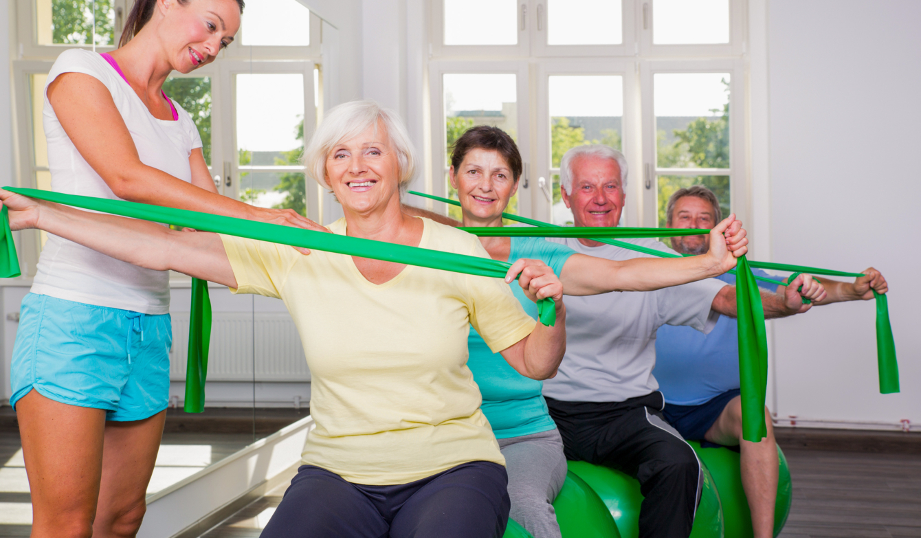 Visualbild Adobe Stock. Vier Seniorinnen und Senioren sitzen auf einem grünen Gymnastikball und stretchen gleichzeitig einen grünen Theraband. Eine Kursleiterin steht links im Bild und korrigiert die Haltung einer Seniorin.