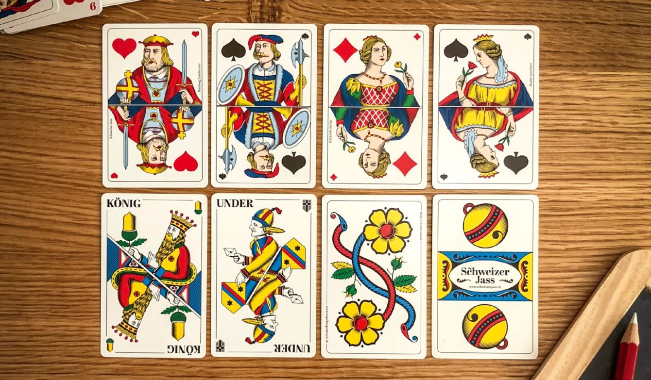 Auf einem Holztisch liegen acht Jasskarten in zwei Reihen. Oben sind französische Bildkarten wie König, Dame und Bube in kräftigen Farben zu sehen. Unten liegen traditionelle Schweizer Jasskarten mit typischen Motiven wie König, Under, einer bunten Blumenkarte und der Rückseite mit dem Schriftzug «Schweizer Jass». Rechts am Rand ist ein Teil eines Holzbretts und zwei rote Bleistifte sichtbar.