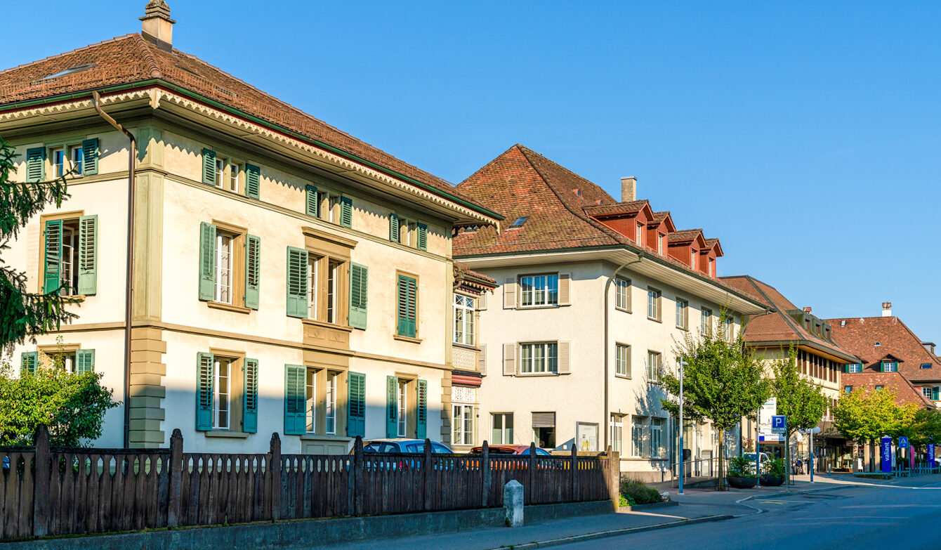 Gebäude der Gemeindeverwaltung von Münchenbuchsee im Jahr 2018. Mehrere traditionelle Verwaltungsgebäude mit Ziegeldach und Fensterläden stehen an einer ruhigen Strasse unter blauem Himmel.
