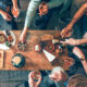 Mehrere Personen sitzen und stehen um einen tiefen Holztisch, auf dem verschiedene Snacks und kleine Speisen wie Brot, Käse, Nüsse, Aufschnitt und Cracker angerichtet sind. Einige Personen halten Gläser mit einem hellen Getränk in der Hand. Die Szene zeigt eine gesellige Tischsituation von oben, mit vielen Händen, die nach Speisen greifen oder Gläser anheben.