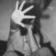 Schwarzweisses Foto in körniger Optik. Im Vordergrund ist eine offene Hand zu sehen, mit gespreizten Fingern, die direkt zur Kamera zeigt und einen grossen Teil des Bildes einnimmt. Die Hand wirkt sehr nah, fast berührend. Im Hintergrund befindet sich eine gebeugte Person, unscharf und teilweise verdeckt durch die erhobene Hand. Der Körper ist zusammengesunken, die Beine sind angezogen, die Knie sichtbar. Die Kleidung zeigt feine Streifen und Punkte. Der Hintergrund ist dunkel und undefiniert, ohne erkennbare Details des Raums. Das Bild vermittelt Distanz und Abwehr durch die nach vorne gerichtete Hand, während der restliche Körper in den Hintergrund zurücktritt.
